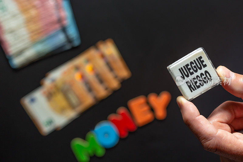 两个成年男性的手指握着一个老虎机游戏按钮，上面写着“juegue Riesgo”(英语:play Risk)。下面是——模糊的——一叠欧元钞票和“钱”这个词。